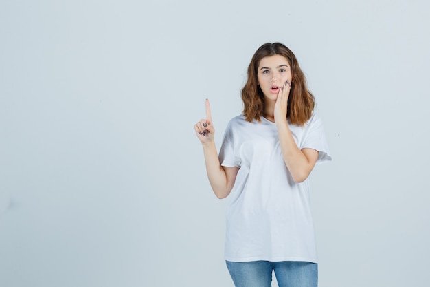 Young lady in t-shirt, jeans pointing up while holding hand on cheek and looking puzzled , front view.