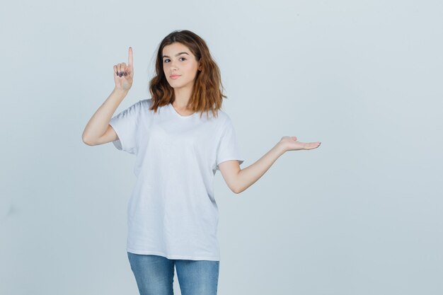 Young lady in t-shirt, jeans pointing up while showing something and looking confident , front view.