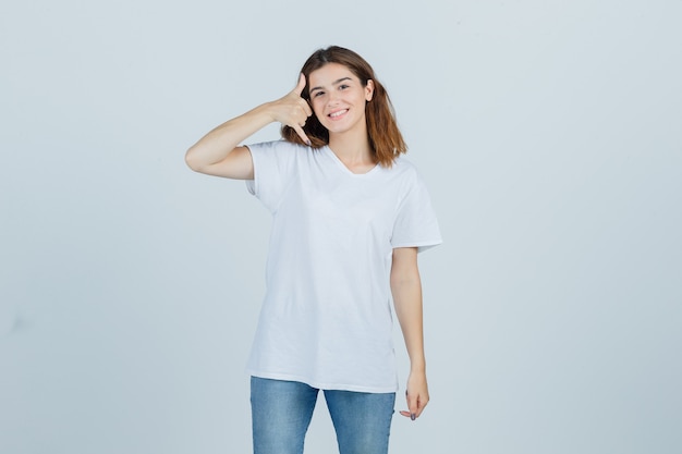 Young lady in t-shirt, jeans showing phone gesture and looking joyful , front view.