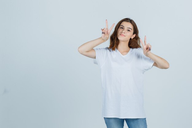 Девушка в футболке, джинсах показывает знак победы и выглядит веселой, вид спереди.