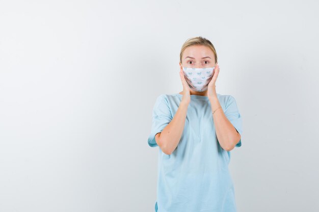 Young lady in t-shirt, mask holding hands on cheeks and looking perplexed