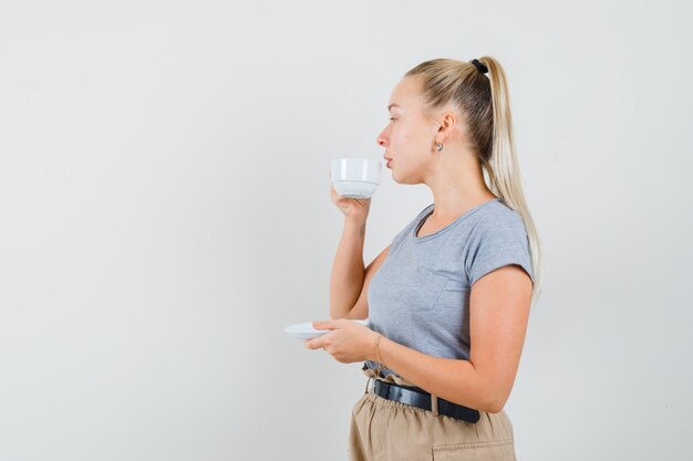 Young lady in t-shirt and pants drinking tea while looking away and looking thoughtful