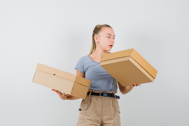 Young lady in t-shirt and pants holding cardboard boxes and looking focused