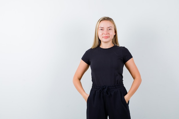 Young lady in t-shirt, pants holding hands in pockets and looking merry , front view.