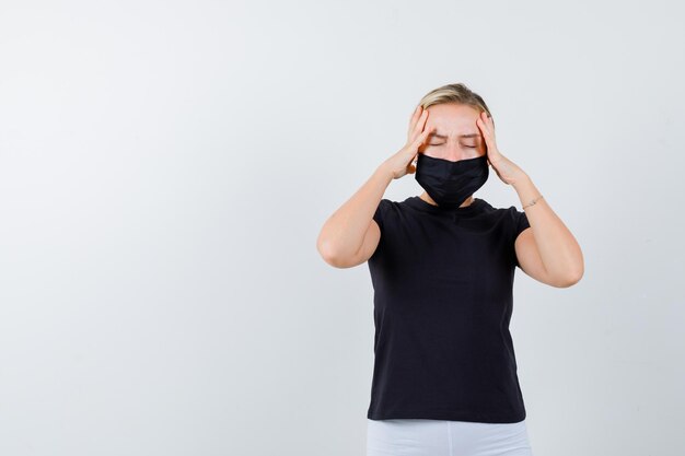 Young lady in t-shirt, pants, medical mask holding hands on head isolated