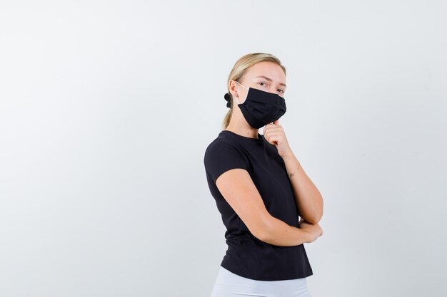 Young lady in t-shirt, pants, medical mask standing in thinking pose isolated