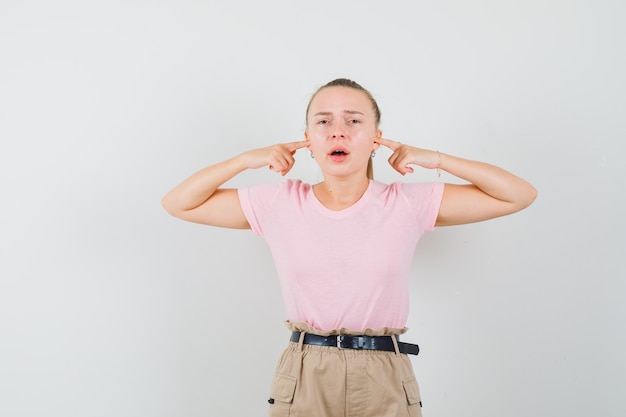 Young lady in t-shirt and pants plugging ears with fingers and looking annoyed