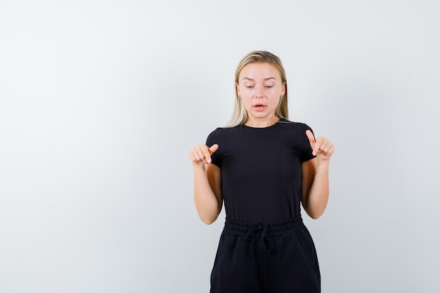 Young lady in t-shirt, pants pointing down and looking pensive , front view.