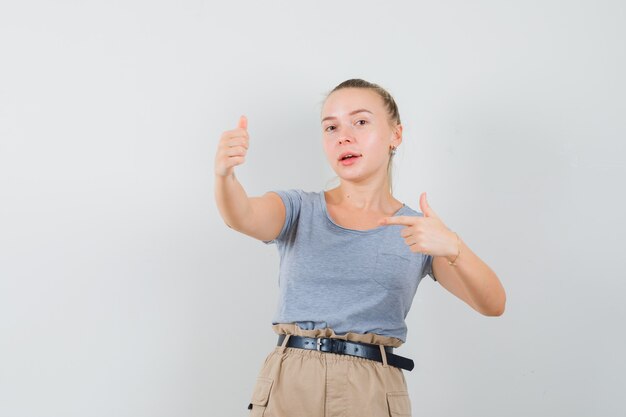 Young lady in t-shirt and pants pointing at her thumb up and looking jovial