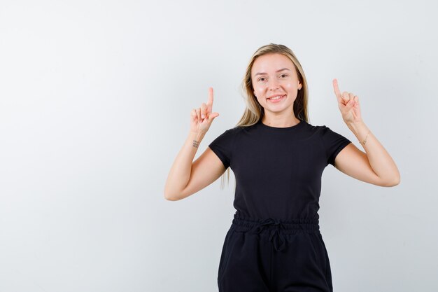 Young lady in t-shirt, pants pointing up and looking joyful , front view.