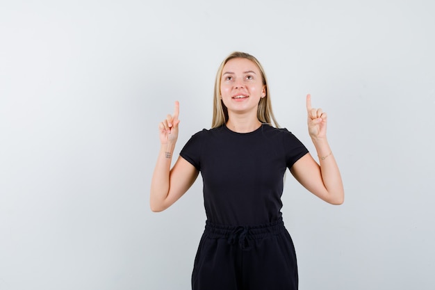 Young lady in t-shirt, pants pointing up and looking merry , front view.