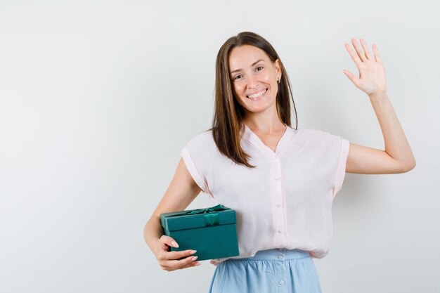 Young lady in t-shirt, skirt holding present box while showing palm and looking joyful , front view.