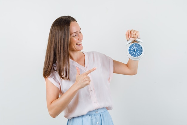 Young lady in t-shirt, skirt pointing at alarm clock and looking joyful , front view.