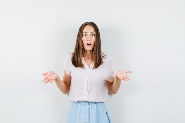 Young lady in t-shirt, skirt raising palms and looking puzzled , front view.