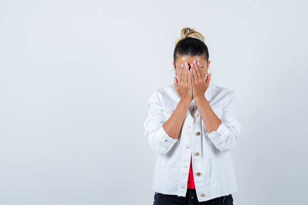 Young lady in t-shirt, white jacket covering face with hands and looking disappointed , front view.