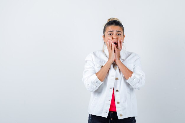 Young lady in t-shirt, white jacket with hands on mouth and looking scared , front view.