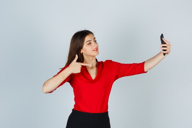 Young lady taking selfie while pointing to the right side in red blouse