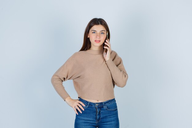 베이지색 스웨터, 청바지를 입고 즐거운 표정으로 휴대폰으로 통화하는 젊은 여성, 전면 전망.