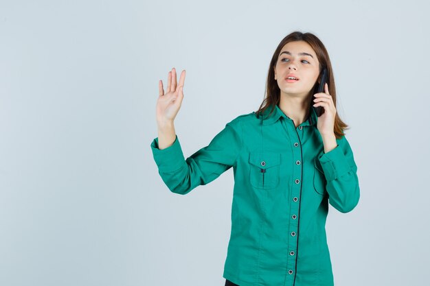 携帯電話で話している若い女性は、緑色のシャツで停止ジェスチャーを示し、自信を持って見えます。正面図。