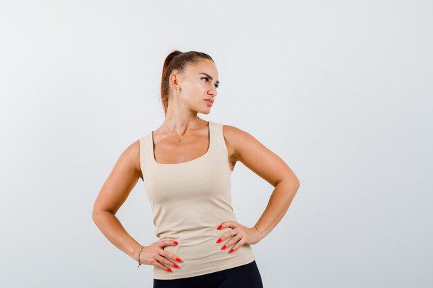 Young lady in tank top keeping hands on waist and looking pensive , front view.