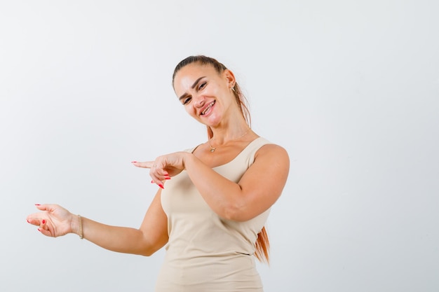 Young lady in tank top pointing aside and looking cheerful, front view.