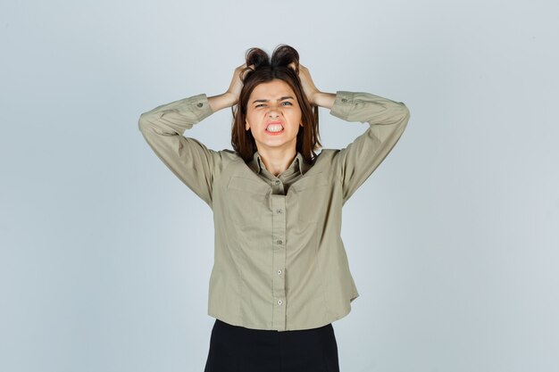 Young lady tearing at hair with her hands while clenching teeth in shirt, skirt and looking aggressive , front view.