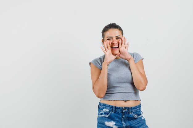 Young lady telling secret with hands near mouth in t-shirt, shorts and looking merry. 