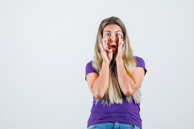 Young lady telling secret with hands near mouth in violet t-shirt, jeans front view.