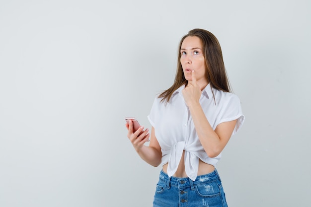 Young lady thinking something while holding device in white blouse and looking thoughtful , front view. space for text