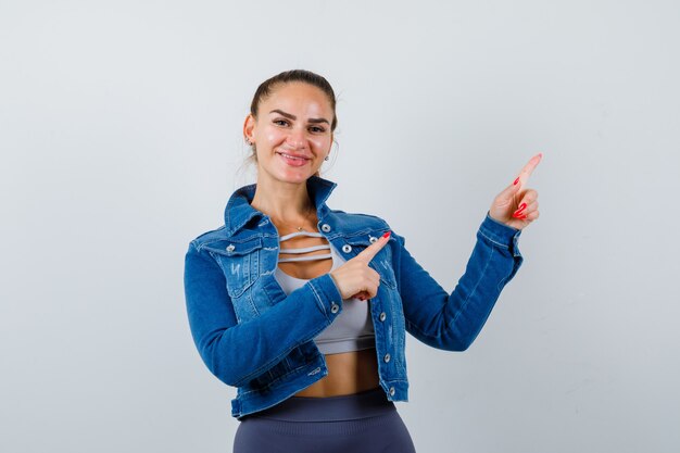 Young lady in top, denim jacket pointing down and looking joyful , front view.