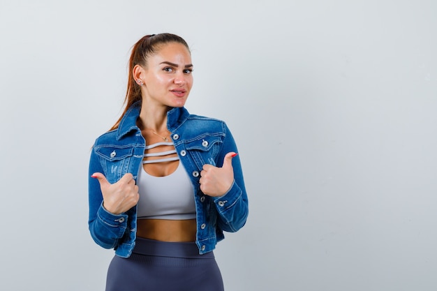 Young lady in top, denim jacket showing thumbs up and looking joyful , front view.