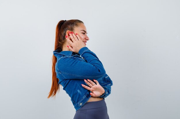 Young lady in top, denim jacket with hand on cheek and looking cheery , front view.