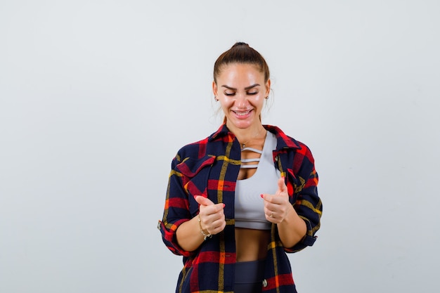 Young lady in top, plaid shirt pretending to hold something and looking merry , front view.