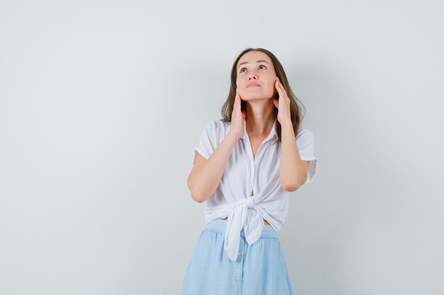 Young lady touching her face with hands in white blouse,blue skirt and looking sensible