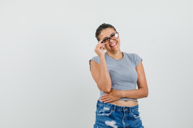 Young lady touching her glasses in t-shirt, shorts and looking cheerful. 