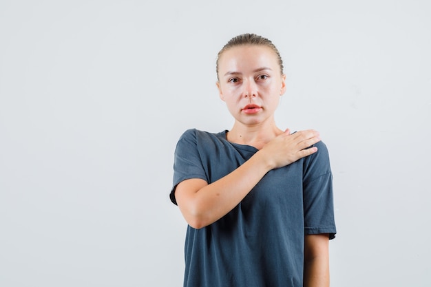 Young lady touching shoulder with hand in grey t-shirt and looking calm