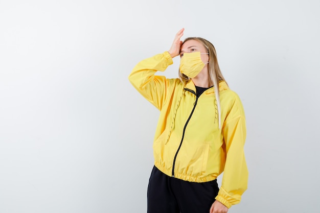 Young lady in tracksuit, mask keeping hand on head and looking fatigued , front view.