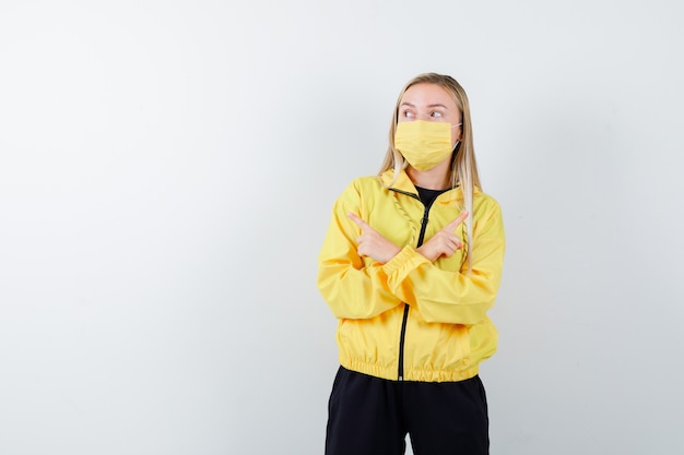 Young lady in tracksuit, mask pointing left and right and looking hesitant , front view.