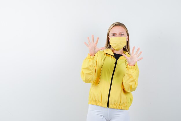 Young lady trying to block herself with hands in jacket, pants, mask and looking terrified , front view.