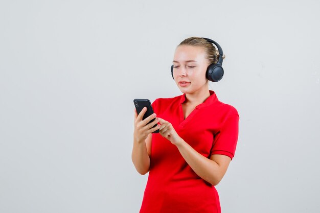 赤いTシャツ、ヘッドフォンで携帯電話に入力し、嬉しそうに見える若い女性