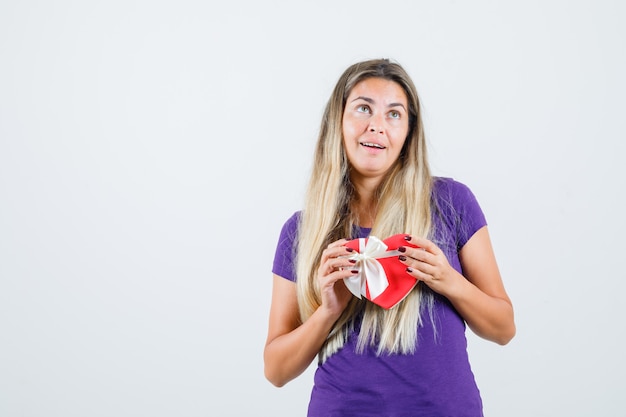 Молодая дама в фиолетовой футболке держит подарочную коробку и выглядит веселой, вид спереди.