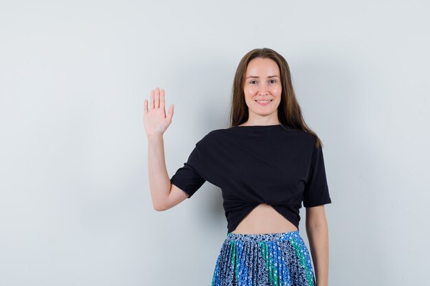 Young lady waving hand in blouse, skirt and looking cheery