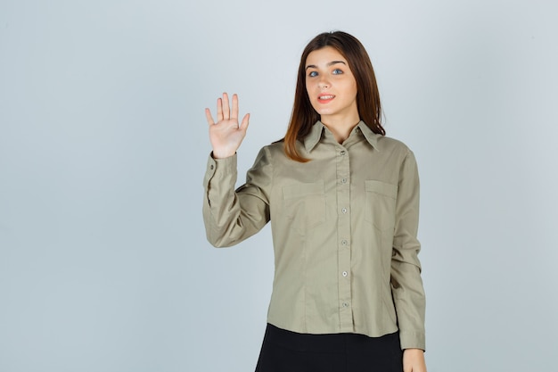 Young lady waving hand for greeting in shirt, skirt and looking cheery. front view.