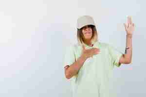 Free photo young lady waving hand for greeting in t-shirt, cap and looking hesitant , front view.