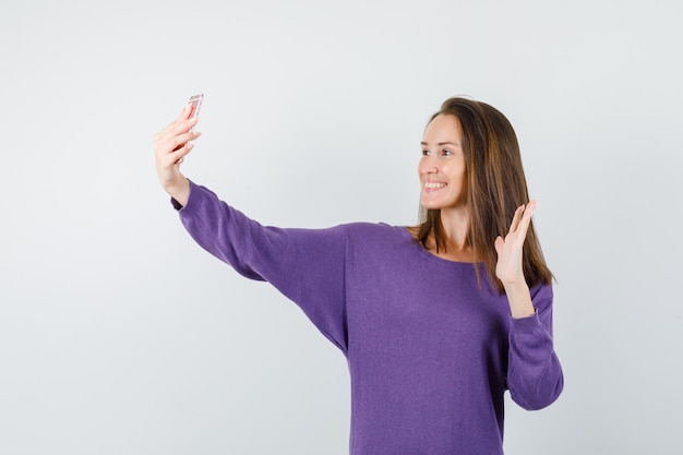 Giovane donna agitando la mano sulla videochiamata in camicia viola e guardando felice. vista frontale.