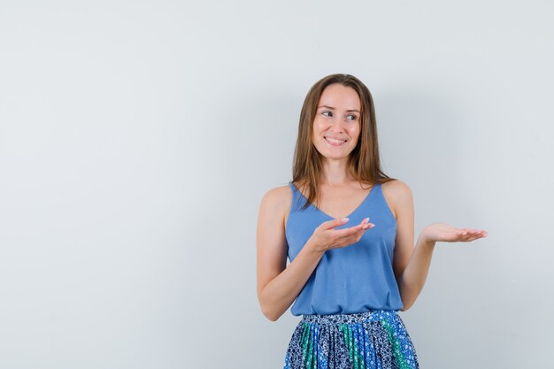 Young lady welcoming or showing something in singlet, skirt and looking cheerful , front view.