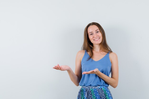 Young lady welcoming or showing something in singlet, skirt and looking cheerful. front view.