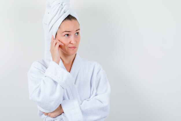 Young lady in white bathrobe, towel holding hand on cheek and looking thougthful , front view.
