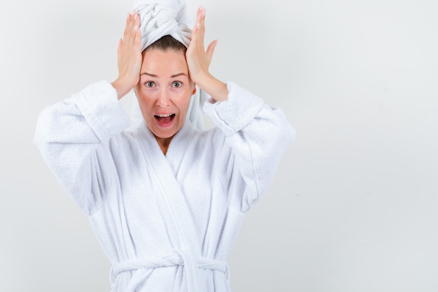 Young lady in white bathrobe, towel holding hands on head and looking distressed , front view.