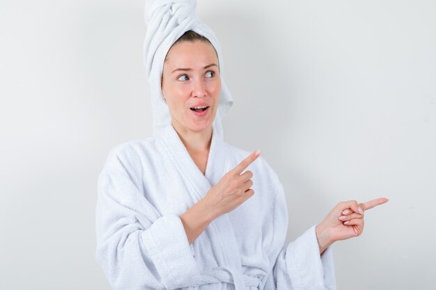 Young lady in white bathrobe, towel pointing aside and looking amazed , front view.